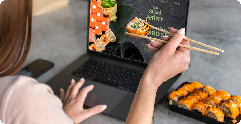 Person using chopsticks to eat sushi while watching a laptop screen showing a sushi website